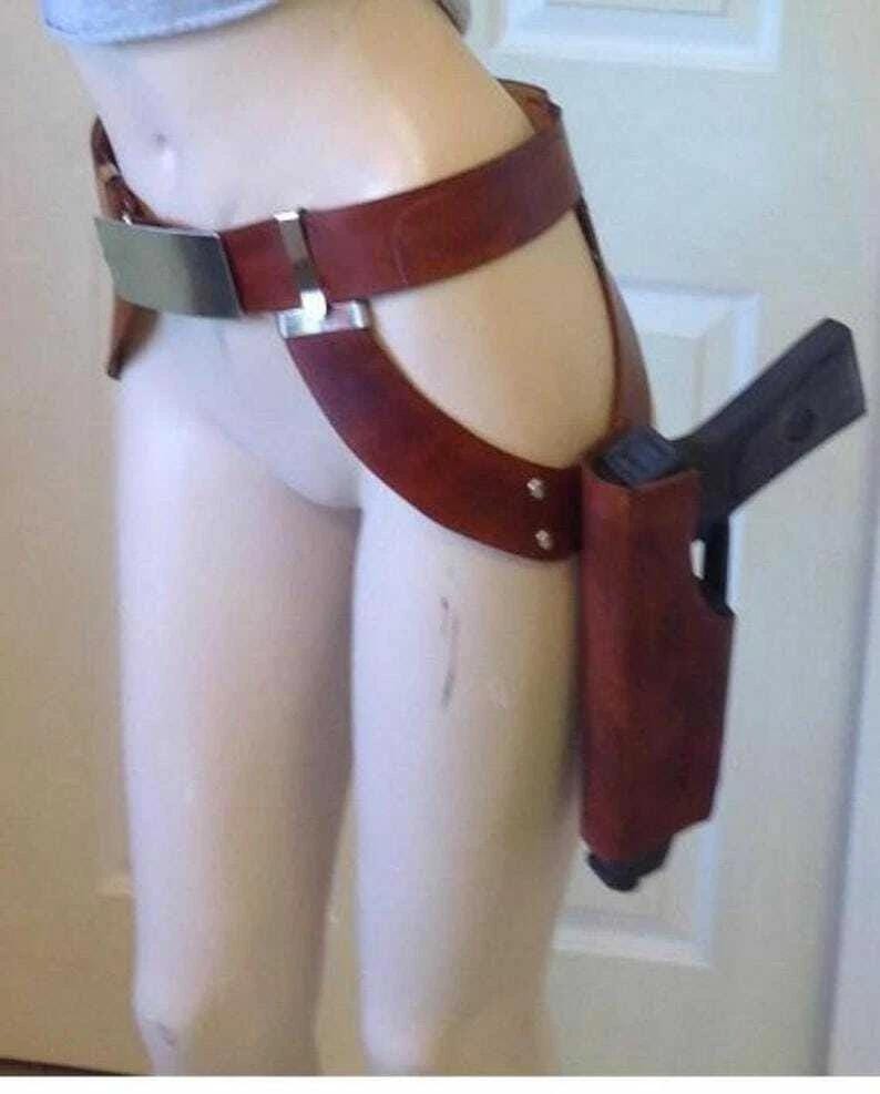 May include: A brown leather thigh holster with a gun. The holster is attached to a belt with a silver buckle and a strap that goes around the thigh. The gun is black.