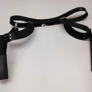 May include: Black leather shoulder holster with two attached pouches. The holster has a belt that wraps around the torso and connects to the shoulder straps. The pouches are designed to hold items, and the holster is secured with metal hardware.