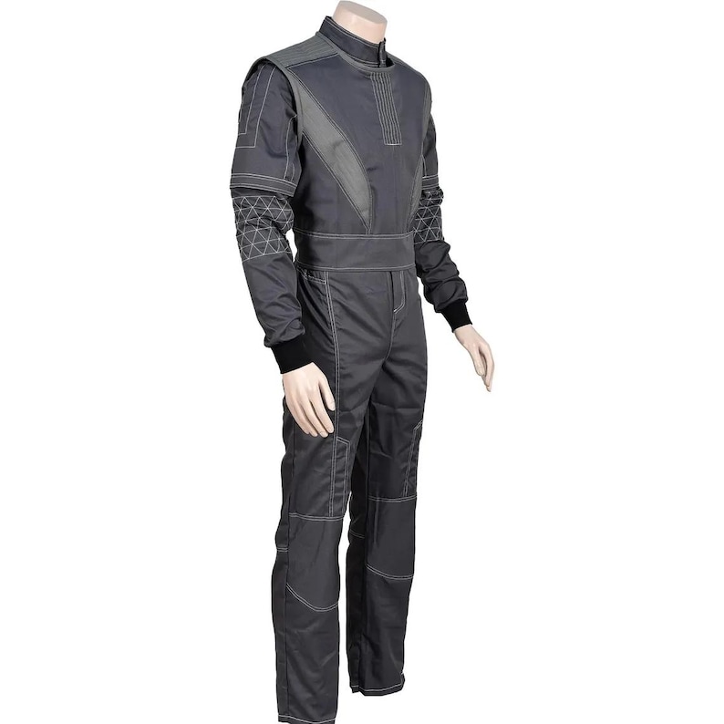 Puede incluir: Mono de carreras gris oscuro con paneles grises en contraste y costuras blancas. El mono presenta un cuello alto, mangas largas con pu&ntilde;os negros y un cintur&oacute;n en la cintura. El dise&ntilde;o incluye patrones geom&eacute;tricos.