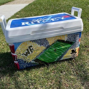 May include: A white and blue cooler with the text "RITCEY" on the lid. The cooler is decorated with a football field, the letters "WF", and a cartoon character. The cooler has white handles.