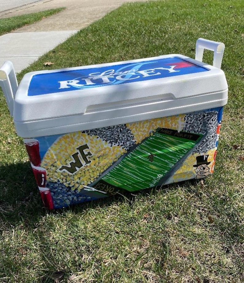 May include: A white and blue cooler with the text "RITCEY" on the lid. The cooler is decorated with a football field, the letters "WF", and a cartoon character. The cooler has white handles.