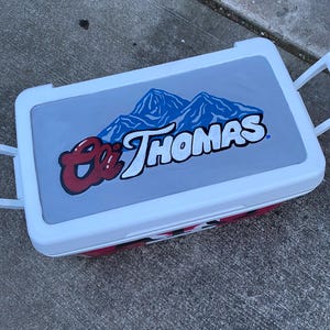 May include: A white and red cooler with a grey lid featuring the text "Eli Thomas" in red and white, with a blue mountain graphic. The cooler has white handles and a red base.