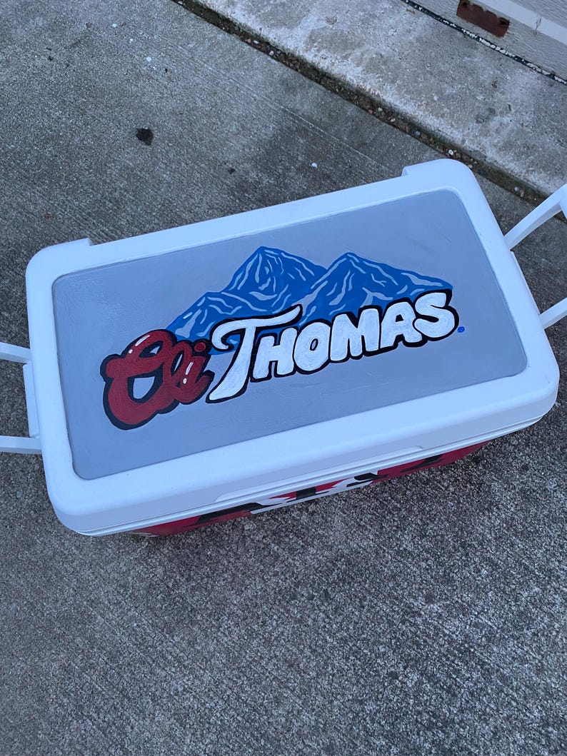 May include: A white and red cooler with a grey lid featuring the text "Eli Thomas" in red and white, with a blue mountain graphic. The cooler has white handles and a red base.