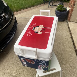 May include: A white and red cooler featuring a Dayton Flyers graphic on the lid. The cooler has a white handle and is placed on a small white stool. The side of the cooler has a navy blue section with the text "Flyers Never Fold".