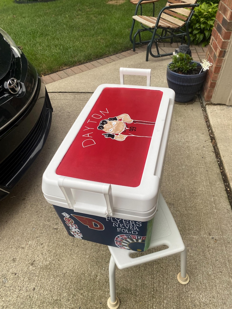 May include: A white and red cooler featuring a Dayton Flyers graphic on the lid. The cooler has a white handle and is placed on a small white stool. The side of the cooler has a navy blue section with the text "Flyers Never Fold".