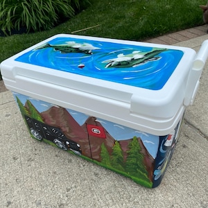 May include: A white cooler with a hand-painted design. The lid shows a blue water scene with fish. The sides depict mountains, trees, and a black vehicle. A red flag is also visible.