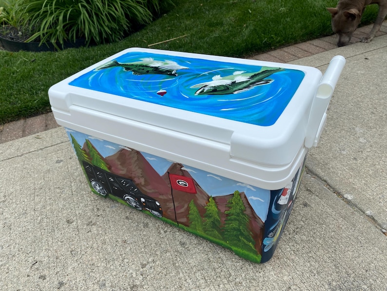 May include: A white cooler with a hand-painted design. The lid shows a blue water scene with fish. The sides depict mountains, trees, and a black vehicle. A red flag is also visible.