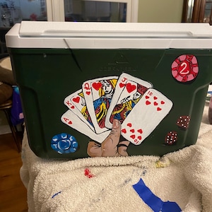 May include: A green cooler with a white lid, painted with playing cards, a hand holding the cards, and a red and white chip with the number 2 on it. The cooler has the text "Ozark Trail" on it.