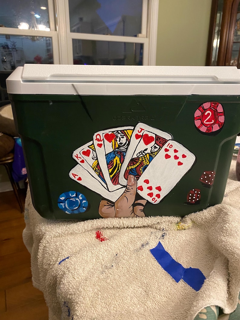 May include: A green cooler with a white lid, painted with playing cards, a hand holding the cards, and a red and white chip with the number 2 on it. The cooler has the text "Ozark Trail" on it.