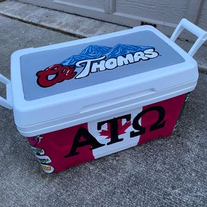 May include: A white and red cooler with a grey lid. The lid features a blue mountain graphic and the text "St. Thomas". The cooler's side displays "AT&Omega;" with a maple leaf design. White handles are on each end.