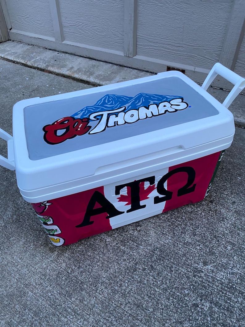 May include: A white and red cooler with a grey lid. The lid features a blue mountain graphic and the text "St. Thomas". The cooler's side displays "AT&Omega;" with a maple leaf design. White handles are on each end.