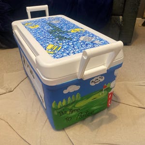 May include: A white and blue cooler featuring a golf course design. The lid is white with a blue and yellow pattern. The side displays the text "It's Time To Partee". The cooler has white handles for easy carrying.