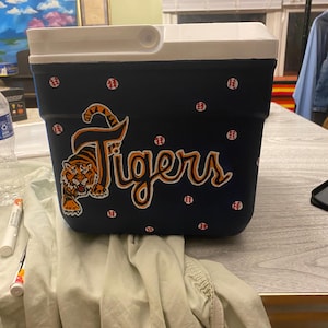 May include: A blue and white cooler with a tiger and the word "Tigers" painted on it. The cooler is decorated with white polka dots and red baseballs.