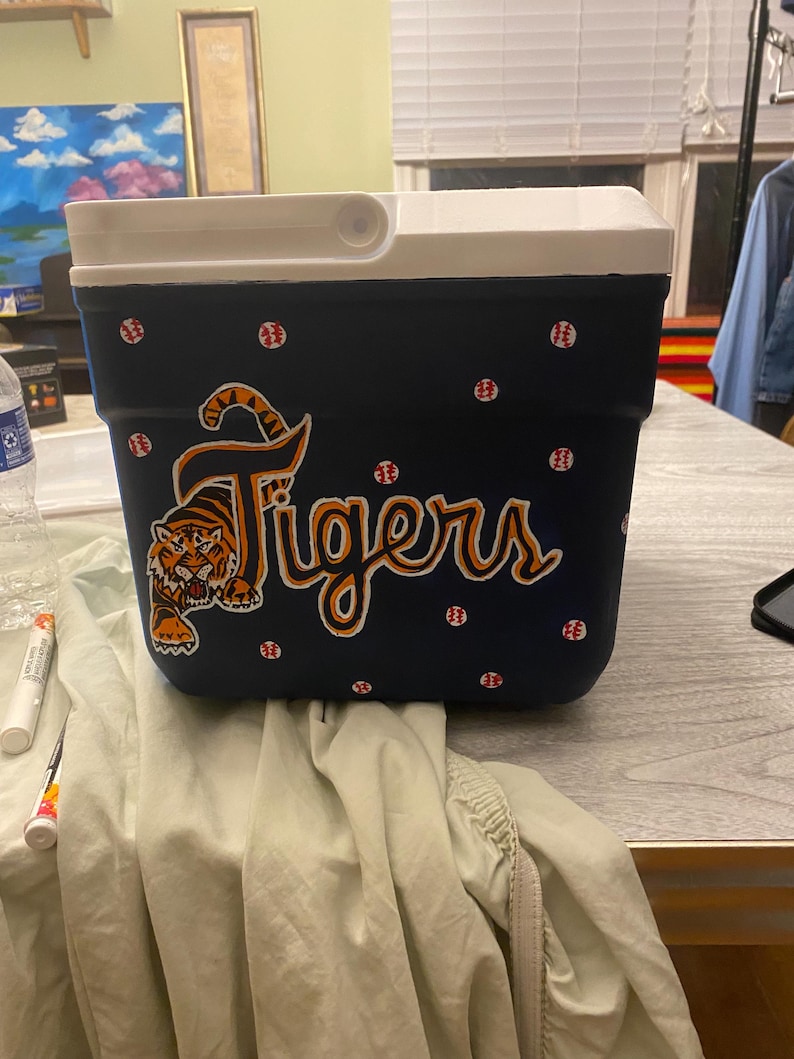 May include: A blue and white cooler with a tiger and the word "Tigers" painted on it. The cooler is decorated with white polka dots and red baseballs.