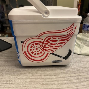 May include: White cooler with a red Detroit Red Wings logo, a black hockey stick, and a black hockey puck. The cooler has a blue stripe around the bottom.
