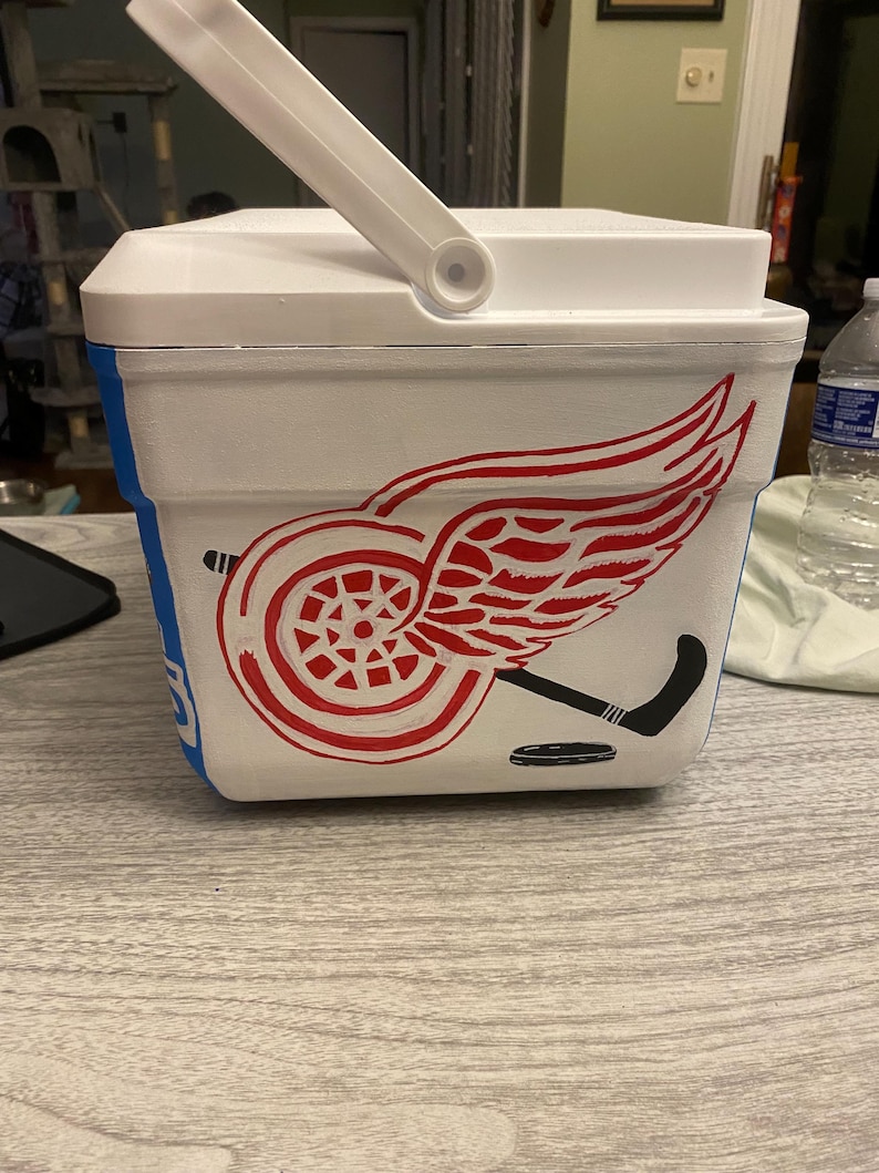 May include: White cooler with a red Detroit Red Wings logo, a black hockey stick, and a black hockey puck. The cooler has a blue stripe around the bottom.