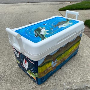May include: White cooler with a hand-painted fishing theme. The lid shows a blue water scene with fish. The sides depict a fisherman, fish, and aquatic elements. Includes white handles for easy carrying.