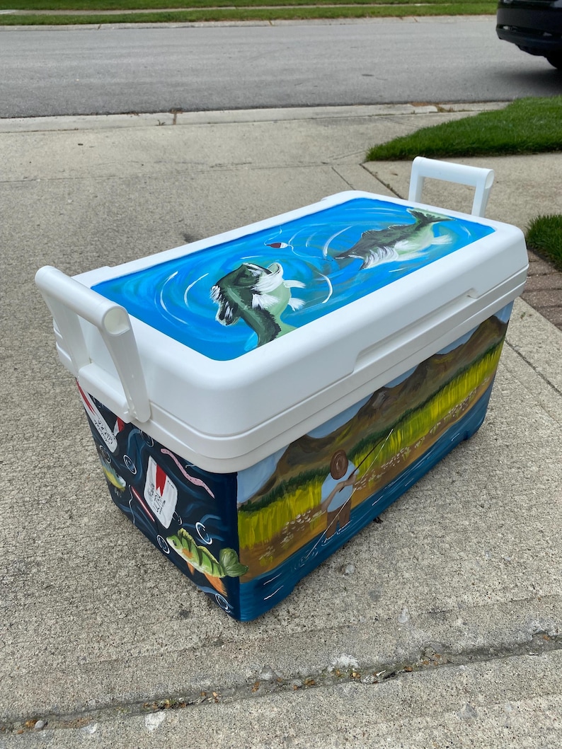 May include: White cooler with a hand-painted fishing theme. The lid shows a blue water scene with fish. The sides depict a fisherman, fish, and aquatic elements. Includes white handles for easy carrying.