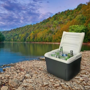 May include: A black and white cooler with a white lid open, revealing a variety of beverages, including bottles and cans, on a bed of ice. The cooler is sitting on a rocky shore with a lake and trees in the background.