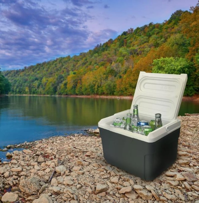 May include: A black and white cooler with a white lid open, revealing a variety of beverages, including bottles and cans, on a bed of ice. The cooler is sitting on a rocky shore with a lake and trees in the background.