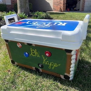 May include: White cooler with a blue and white lid. The cooler is painted with a green pool table design, complete with billiard balls and the words "Chi Kappa" in cursive. White handles are on each side.