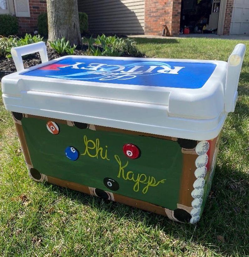 May include: White cooler with a blue and white lid. The cooler is painted with a green pool table design, complete with billiard balls and the words "Chi Kappa" in cursive. White handles are on each side.