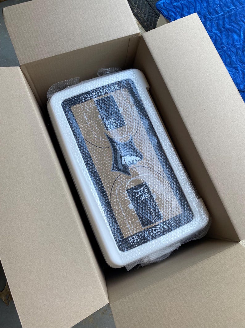 May include: A white and black basketball backboard with the text "PROVIDENCE" and "BIG EAST" inside a cardboard box. The backboard is wrapped in bubble wrap.