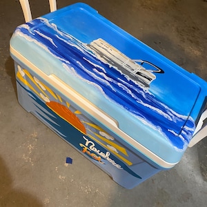 May include: A blue cooler with a white handle and a painted ocean scene. The artwork includes a boat, waves, a sunset, and the word "Nowhere" in cursive. The cooler is designed for outdoor use and has a white handle.