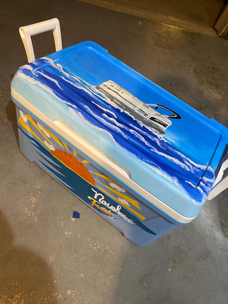 May include: A blue cooler with a white handle and a painted ocean scene. The artwork includes a boat, waves, a sunset, and the word "Nowhere" in cursive. The cooler is designed for outdoor use and has a white handle.