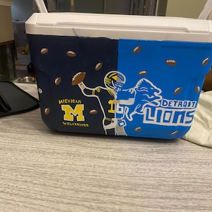 May include: A white cooler with a blue and navy blue design. One side features a football player in a blue and yellow uniform with the text "MICHIGAN WOLVERINES". The other side features a blue lion with the text "DETROIT LIONS".