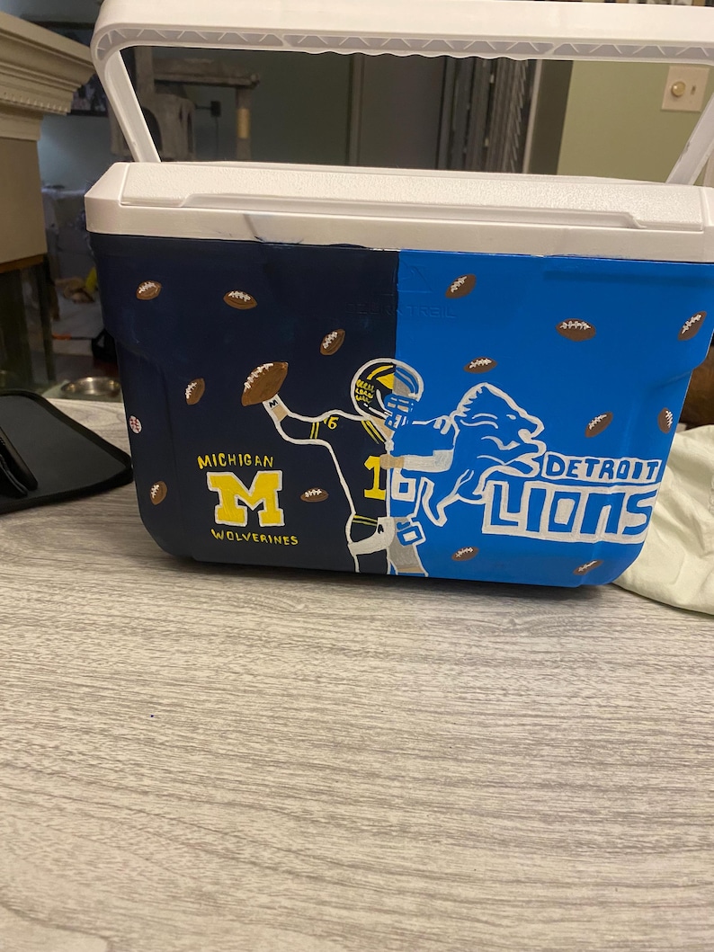 May include: A white cooler with a blue and navy blue design. One side features a football player in a blue and yellow uniform with the text "MICHIGAN WOLVERINES". The other side features a blue lion with the text "DETROIT LIONS".