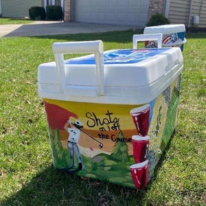 May include: White cooler with a hand-painted golf-themed design. The cooler features a golfer, red cups, and the text "Shots on 3 off the Course." The cooler has a white handle and a blue lid.