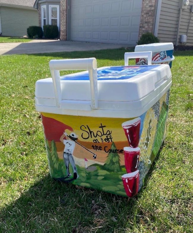 May include: White cooler with a hand-painted golf-themed design. The cooler features a golfer, red cups, and the text "Shots on 3 off the Course." The cooler has a white handle and a blue lid.