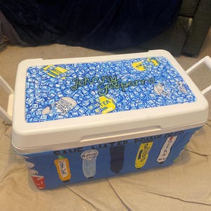 May include: A white and blue cooler with a hand-painted design. The lid features a blue background with ice cubes and the text "Johnny Gapparo Tea". The cooler's side has painted beverage logos.