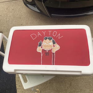 May include: A white cooler with a red lid. The lid displays the word "DAYTON" in white script and a cartoon figure wearing a jersey with the number "99".