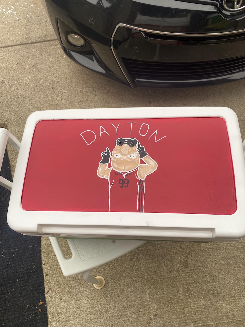 May include: A white cooler with a red lid. The lid displays the word "DAYTON" in white script and a cartoon figure wearing a jersey with the number "99".