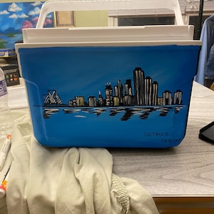 May include: A blue cooler with a hand-painted black and white cityscape of Detroit, Michigan. The cooler has a white lid and a white handle. The words "Detroit Skyline" are written in black on the cooler.