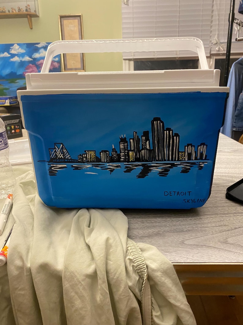 May include: A blue cooler with a hand-painted black and white cityscape of Detroit, Michigan. The cooler has a white lid and a white handle. The words "Detroit Skyline" are written in black on the cooler.