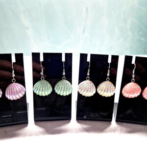 May include: Four pairs of seashell earrings in iridescent pink, green, cream, and coral colours. Each pair is displayed on a black stand with silver-coloured ear wires. The earrings have a textured, shell-like appearance.