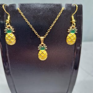 May include: A gold-coloured jewellery set featuring a pineapple design. The set includes a necklace with a pendant, and a pair of earrings. The pineapples are yellow with green tops and are set against a gold-coloured metal.