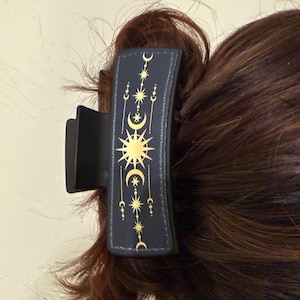 以下が含まれることがあります： 黒いヘアクリップ、天体のデザイン。 クリップは、金色の太陽、月、星で飾られた濃い青色の長方形のパネルが特徴です。 クリップは茶色の髪の一部を保持しています。
