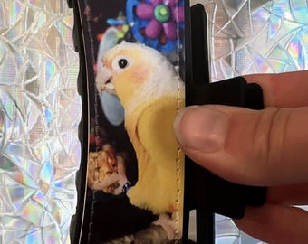 May include: A black hair claw clip featuring a printed design of a yellow and white parrot perched on a branch. The parrot is positioned against a dark background with a colorful blue and purple flower graphic visible behind the bird's head.