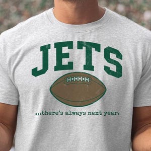 May include: A light gray t-shirt with the word "JETS" in green block letters above a brown football graphic. Below the football, the text reads "...there's always next year."