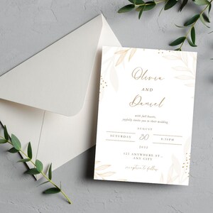 Könnte beinhalten: Eine weiße Hochzeitseinladung mit den Namen "Olivia und Daniel" in goldener Schrift, verziert mit goldenen Blättern. Die Einladung enthält Datum, Uhrzeit und Adresse. Ein Umschlag ist im Hintergrund zu sehen.