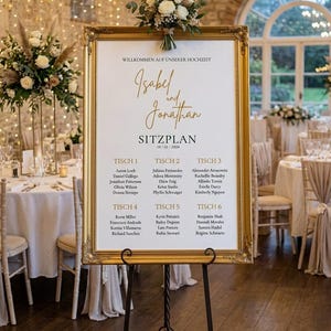 Könnte beinhalten: Ein goldgerahmter Sitzplan für eine Hochzeit, präsentiert auf einer schwarzen Metallstaffelei. Der Plan zeigt die Namen "Isabel und Jonathan" und die Sitzordnung der Gäste. Blumenschmuck und dekorative Beleuchtung unterstreichen das elegante Ambiente.