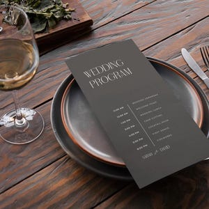 Könnte beinhalten: Ein dunkelgraues Hochzeitsprogramm auf einem Teller mit Gabel und Messer. Das Programm listet den Hochzeitsablauf in weißem Text auf. Ein Glas Weißwein steht auf einem Holztisch.