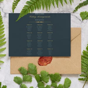 May include: A navy blue wedding seating arrangement card with gold lettering, placed on a kraft paper envelope. The card is surrounded by green fern leaves and a wax seal. The text reads "Ross & Carol Seating Arrangements Wedding Dinner".