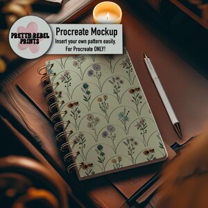 Könnte beinhalten: Ein Spiralnotizbuch mit floralem Muster in Pastellfarben, einer goldenen Spirale und dem Text "Procreate Mockup". Ein Stift und eine brennende Kerze sind ebenfalls zu sehen. Das Notizbuch liegt auf einer braunen Oberfläche.