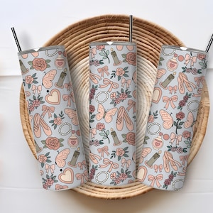 May include: Three stainless steel tumblers with a light blue background and a repeating pattern of ballet shoes, bows, roses, butterflies, and perfume bottles. Each tumbler has a black straw and a silver lid. The tumblers are displayed on a woven tray.