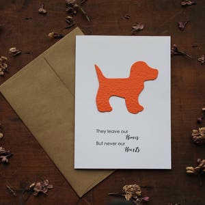May include: A white card with an orange paper cutout of a dog. The card reads "They leave our Homes But never our Hearts".